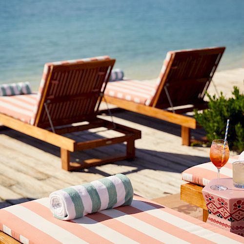Two wooden lounge chairs with striped cushions and towels face a calm body of water, with two drinks placed on a nearby side table.
