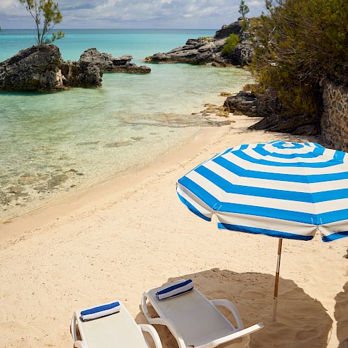 Two lounge chairs with blue towels and a blue-and-white striped umbrella are on a sandy beach by clear water with rocky formations.