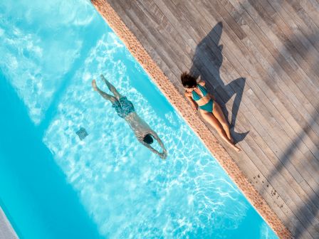 Aerial view of a swimming pool with a person swimming and another sunbathing on the poolside deck, casting a shark-shaped shadow on the deck.