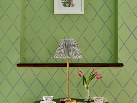 A round table with a lamp, two teacups, and a vase with tulips is placed against a green patterned wall with framed art above it.