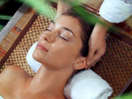 A woman is lying on a spa mat with her eyes closed, receiving a head massage, surrounded by greenery.