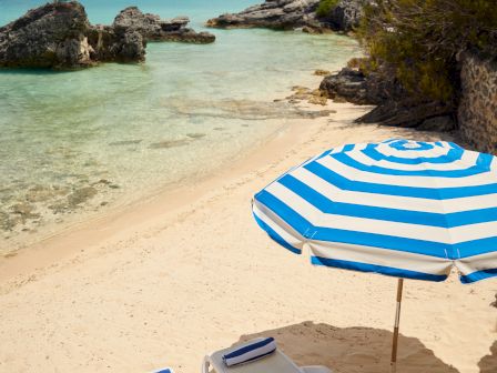 The image shows a beach with two lounge chairs under a blue and white striped umbrella, facing a serene turquoise sea with rocky outcrops.