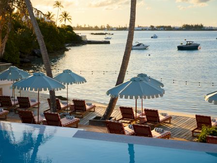 A serene beachfront scene with lounge chairs, striped towels, umbrellas, palm trees, a pool and boats on the water at sunset, all very tranquil.