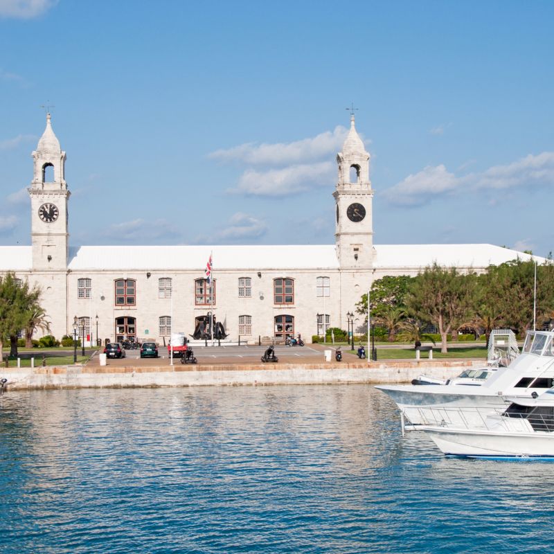 This image shows a building with twin towers and clocks, surrounded by trees and boats docked along the waterfront on a sunny day.