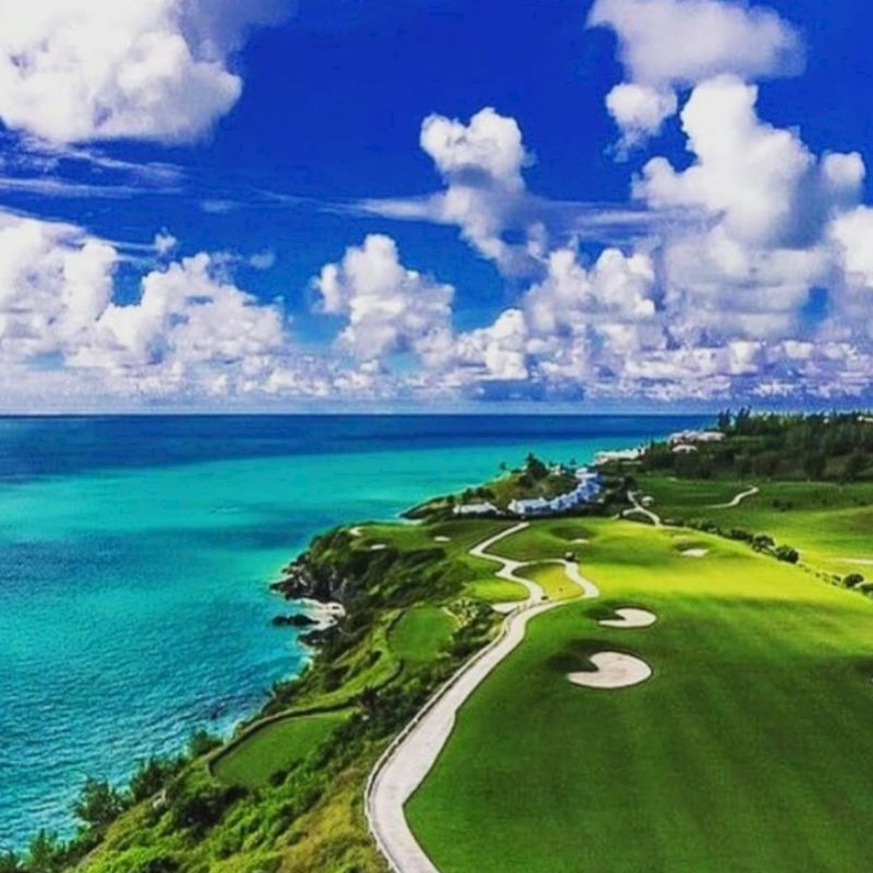 A scenic view of a coastal golf course with a turquoise ocean, green fairways, sand bunkers, and a winding walking path under a partly cloudy sky.