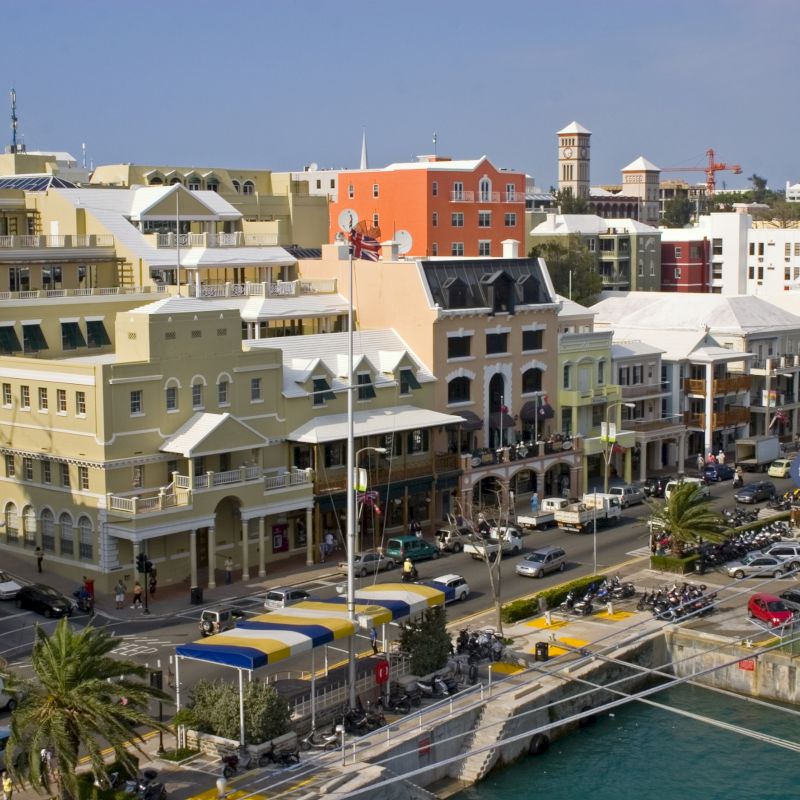 The image shows a coastal cityscape with multiple colorful buildings, street traffic, and docked boats by the waterfront on a sunny day.