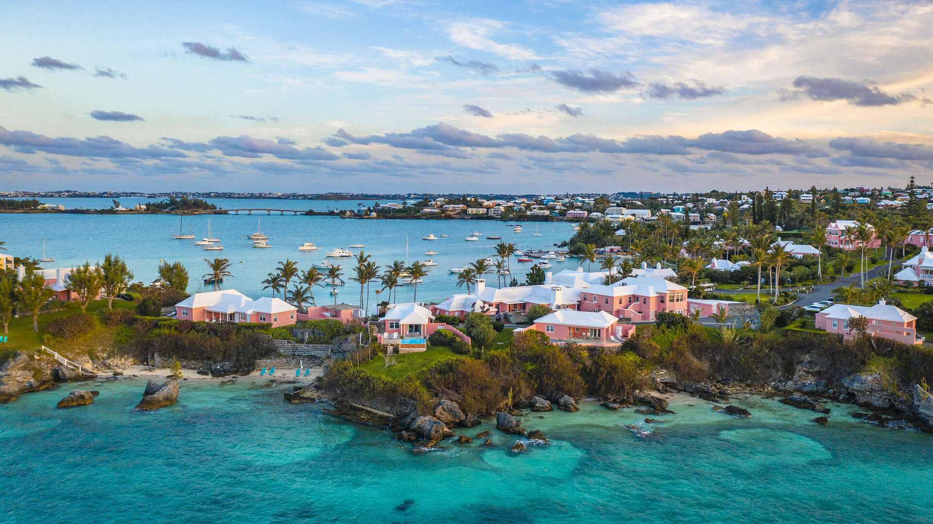 An aerial view of a coastal town featuring pastel-colored buildings, turquoise waters, and boats in the harbor during a partly cloudy day.
