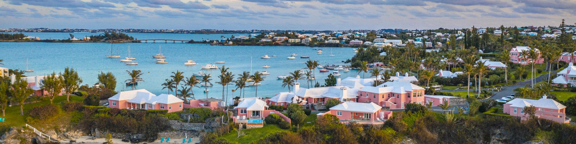 An aerial view of a coastal town featuring pastel-colored buildings, turquoise waters, and boats in the harbor during a partly cloudy day.