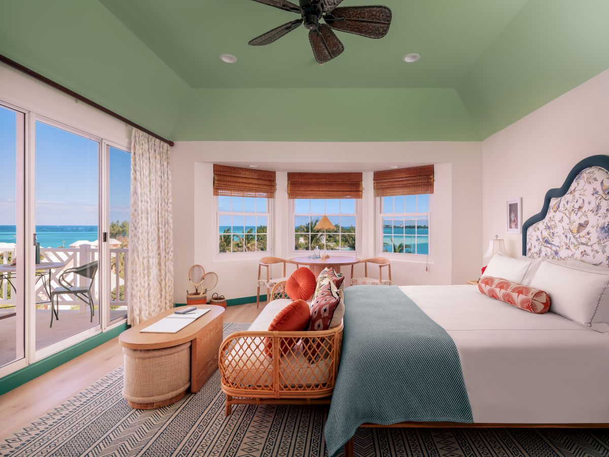 A cozy beachside bedroom with ocean views, large windows, a balcony, ceiling fan, and rattan furniture, with colorful decor and a serene ambiance.
