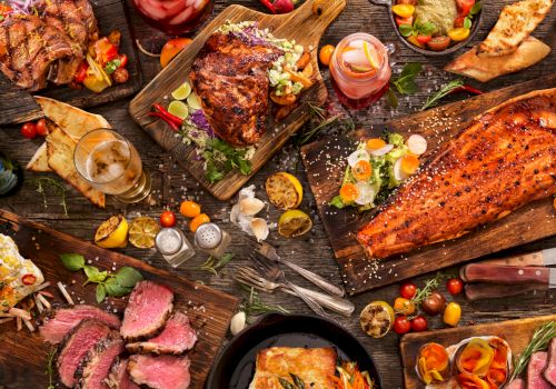 The image features a lavish spread of various dishes including meats, fish, salads, and drinks arranged on a rustic wooden table.