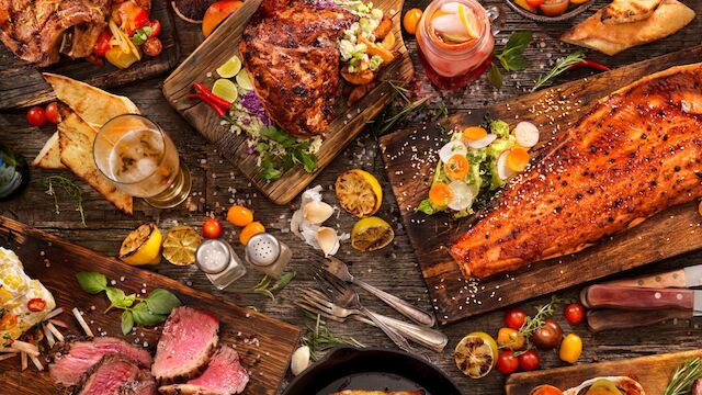 The image features a lavish spread of various dishes including meats, fish, salads, and drinks arranged on a rustic wooden table.