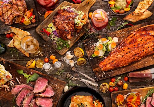 A spread of grilled meats, roasted veggies, and sides on a rustic table; steak, ribs, salmon, and colorful garnishes, plus sauces and bread.
