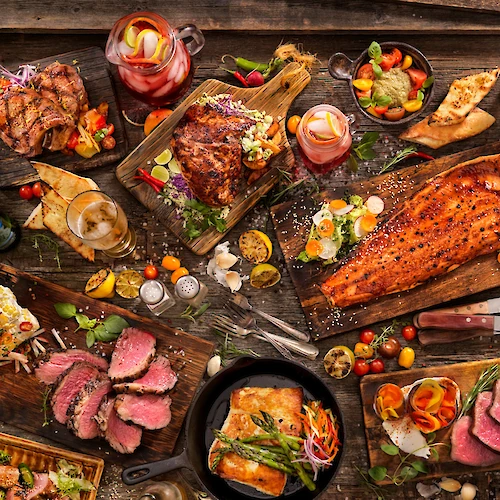 A spread of grilled meats, roasted veggies, and sides on a rustic table; steak, ribs, salmon, and colorful garnishes, plus sauces and bread.