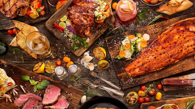 A spread of grilled meats, roasted veggies, and sides on a rustic table; steak, ribs, salmon, and colorful garnishes, plus sauces and bread.