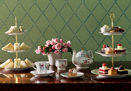 An elegant tea setup with tiered trays of sandwiches and desserts, a teapot, teacups, and a vase of pink flowers on a wooden table.