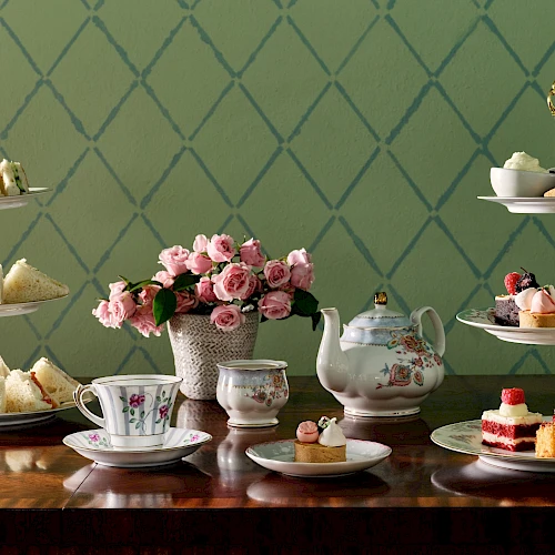 An elegant tea setup with tiered trays of sandwiches and desserts, a teapot, teacups, and a vase of pink flowers on a wooden table.