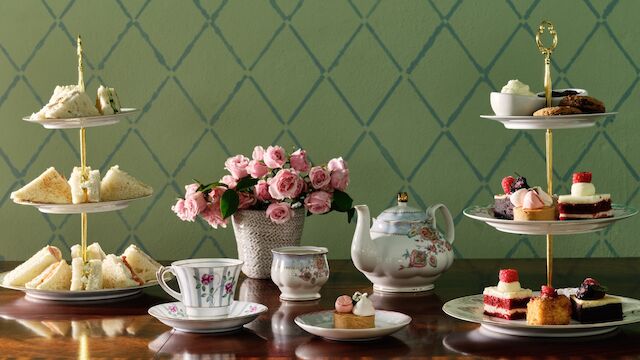 An elegant tea setup with tiered trays of sandwiches and desserts, a teapot, teacups, and a vase of pink flowers on a wooden table.