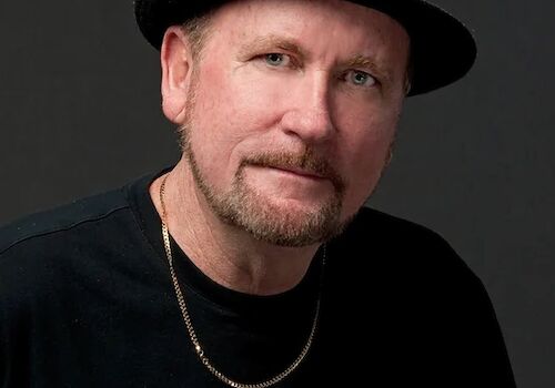 A man wearing a black hat, a black shirt, and a necklace, looking at the camera with a neutral expression, against a dark background.