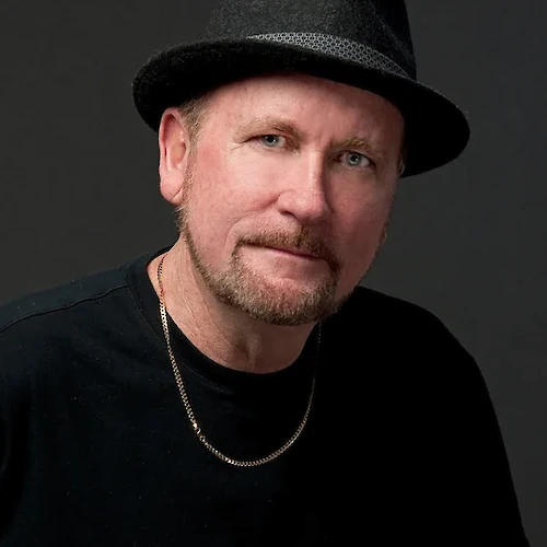 A man wearing a black hat, a black shirt, and a necklace, looking at the camera with a neutral expression, against a dark background.