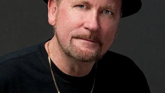 A man wearing a black hat, a black shirt, and a necklace, looking at the camera with a neutral expression, against a dark background.
