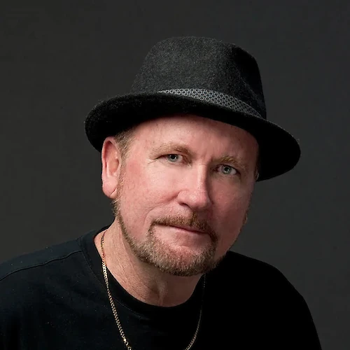 A man wearing a black shirt and a black fedora hat, with a gold chain, against a dark backdrop.