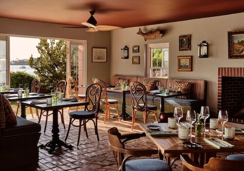 A cozy restaurant interior with wooden tables, comfy chairs, wine glasses, and natural light from the windows. Top it at 140 characters, ending the sentence.