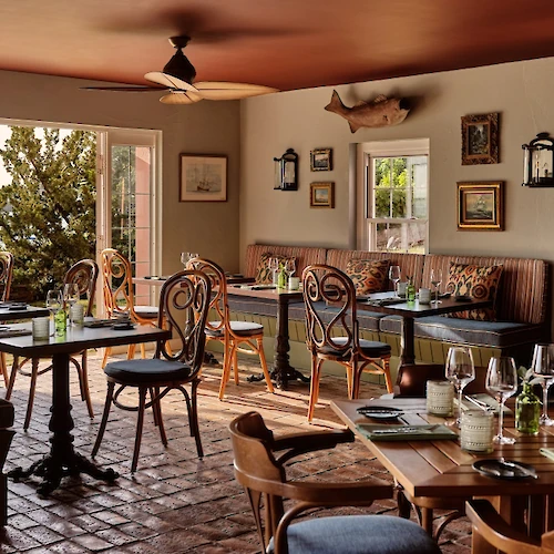 A cozy restaurant interior with wooden tables, comfy chairs, wine glasses, and natural light from the windows. Top it at 140 characters, ending the sentence.