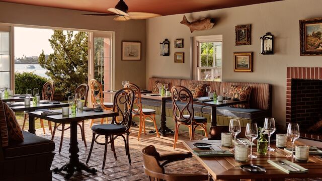 A cozy restaurant interior with wooden tables, comfy chairs, wine glasses, and natural light from the windows. Top it at 140 characters, ending the sentence.