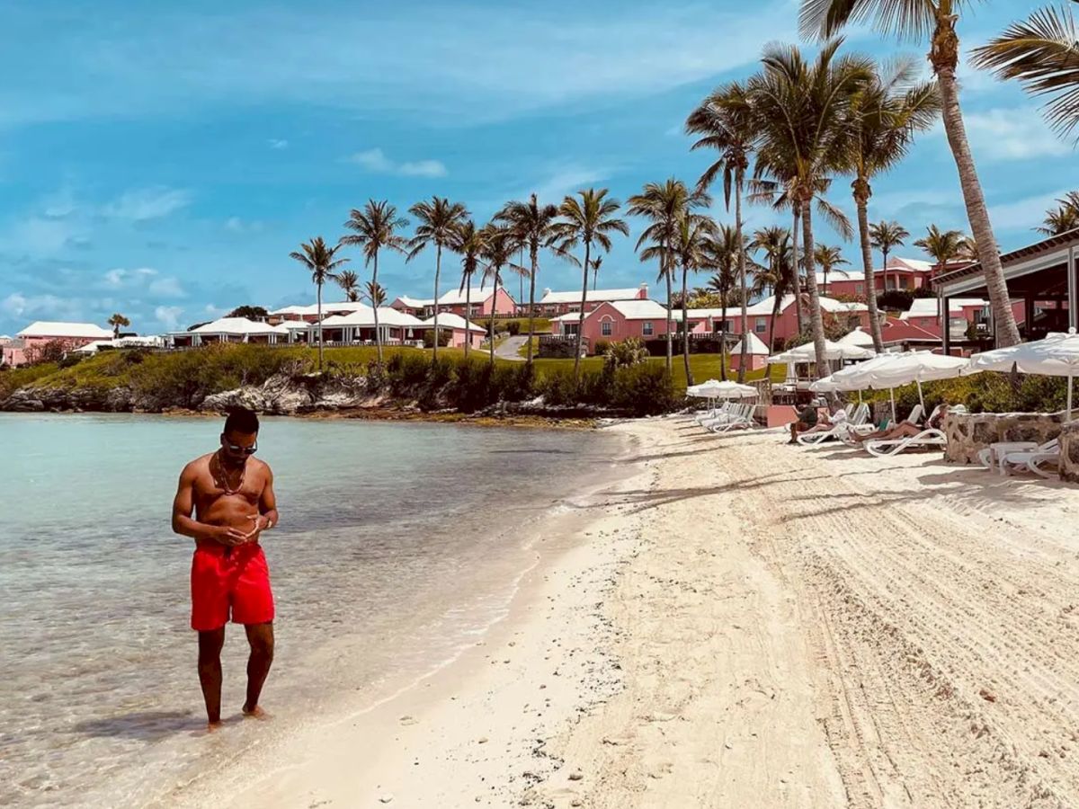 A person in red shorts walks along a sandy beach with palm trees, beach chairs, and umbrellas, adjacent to several buildings under a clear blue sky.