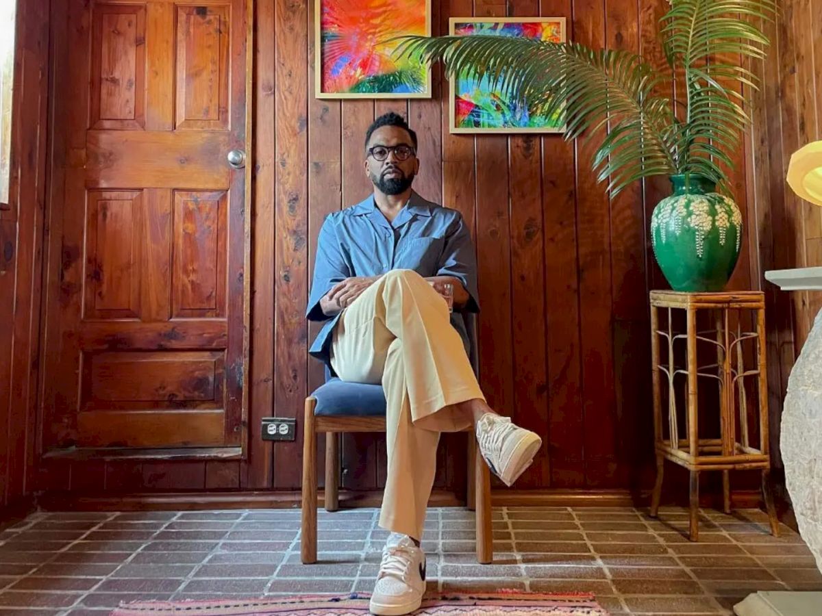 A person is sitting on a chair in a wooden room with plants, artwork, and a lamp, creating a cozy, artistic atmosphere.