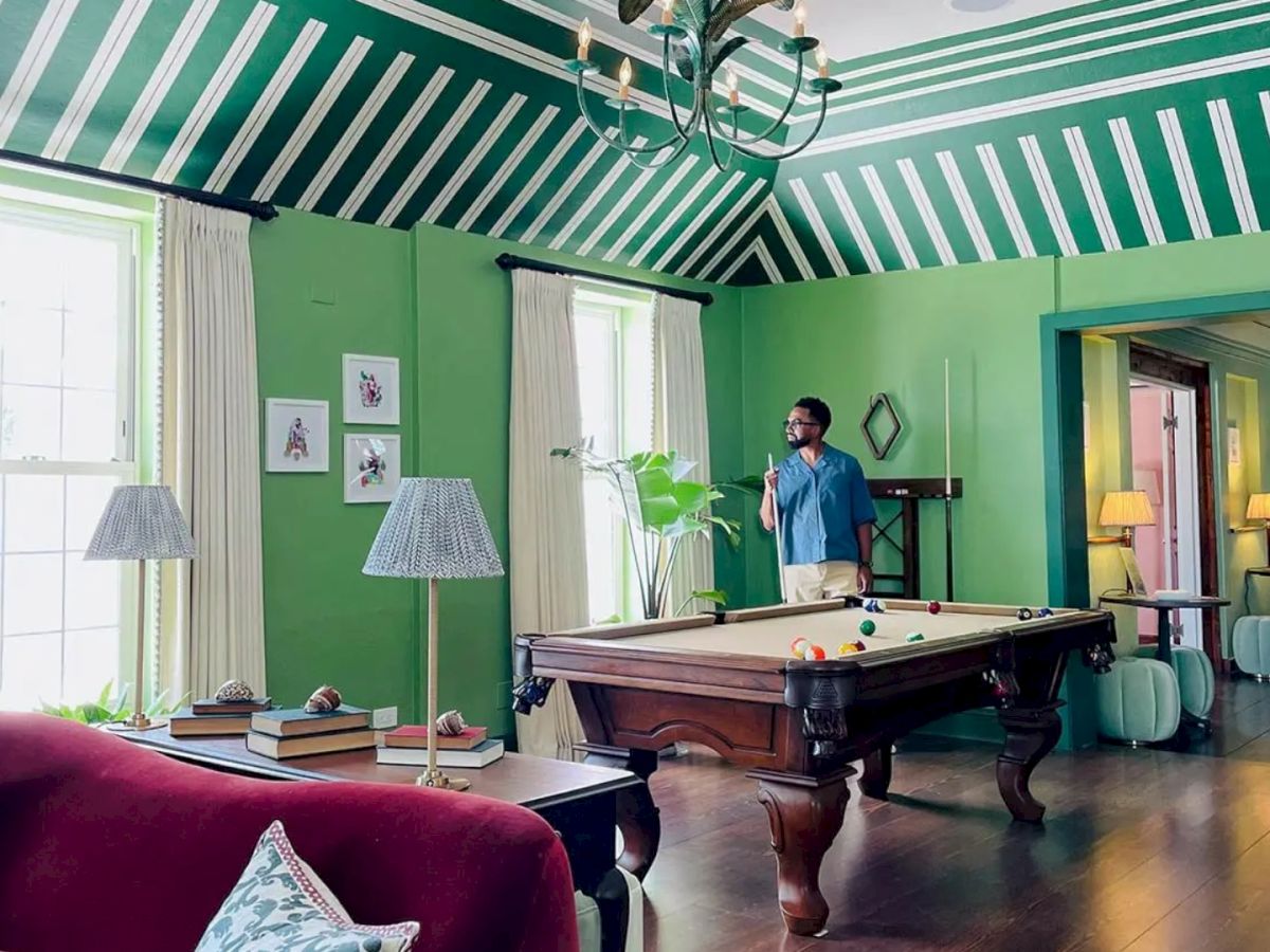 A man stands next to a billiards table in a vibrant green room with striped ceilings, surrounded by furniture, lamps, and framed pictures on the walls.