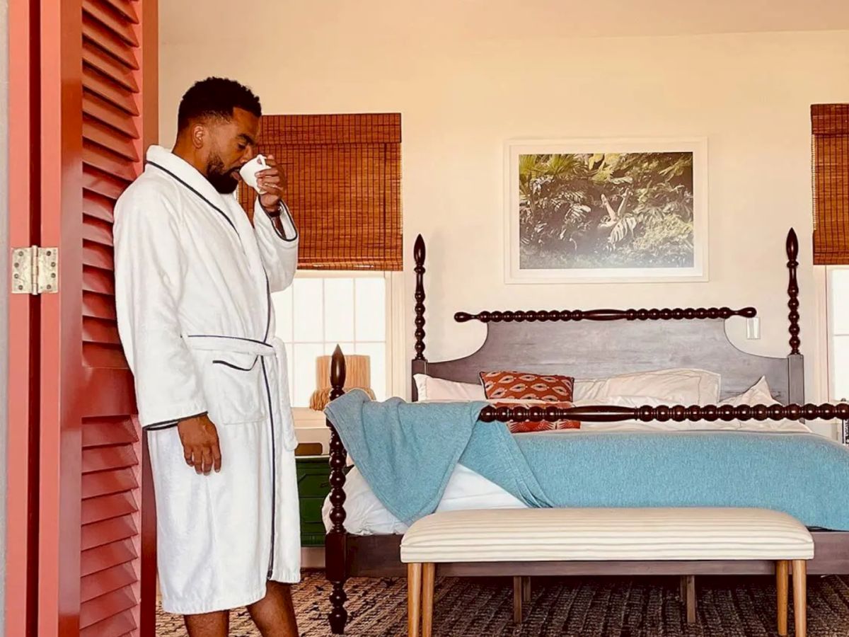 A person in a white robe stands at the foot of a bed in a cozy bedroom, holding a cup, with red shutters and a blanket on the bed.