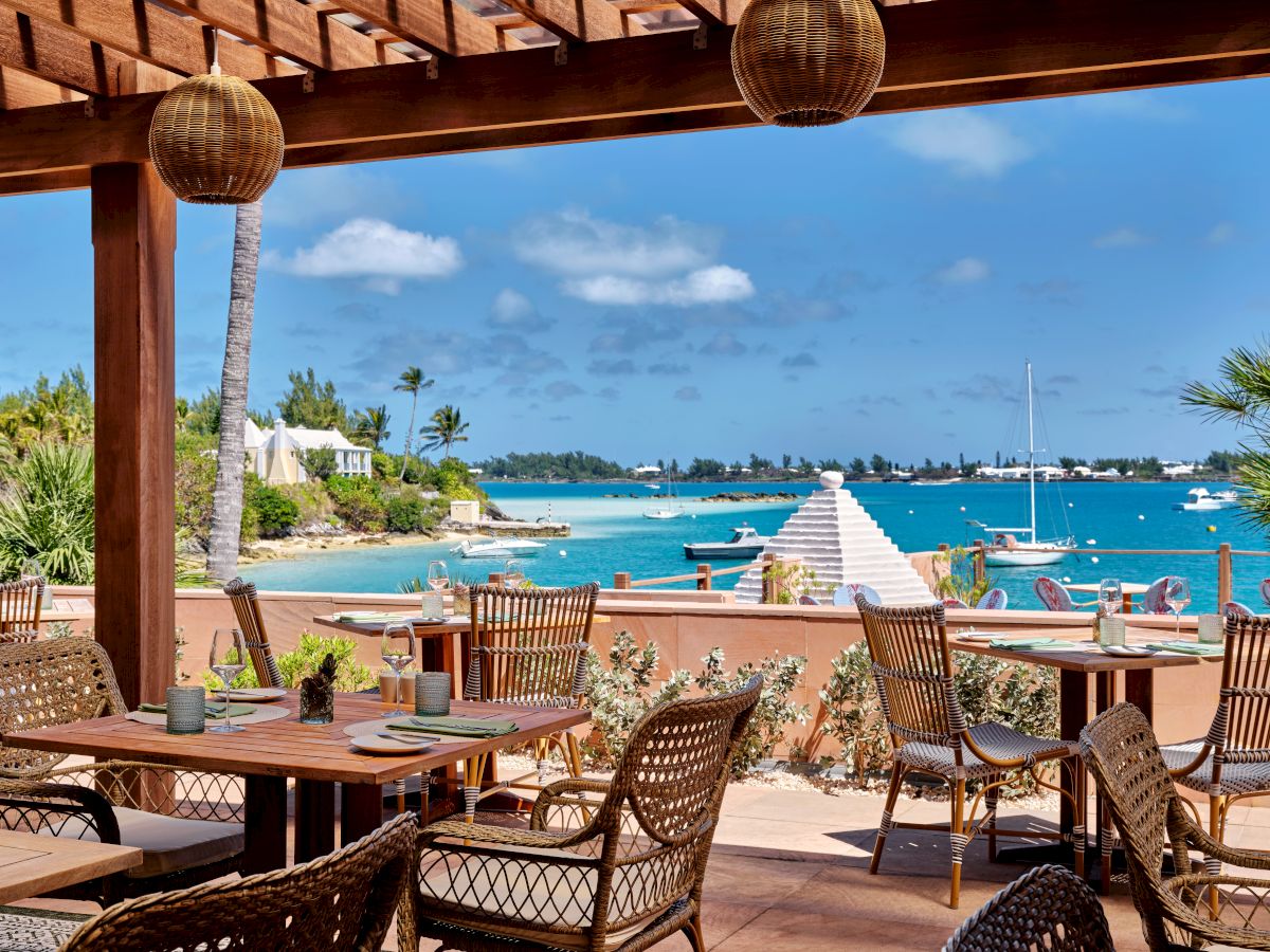 An outdoor dining area with wooden tables and wicker chairs overlooks a scenic coastal view with boats and clear blue water under a sunny sky.