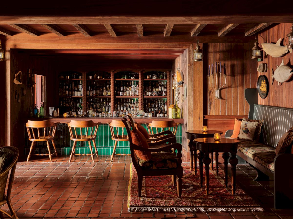 A cozy, rustic bar with wooden furniture, shelves of bottles in the back, a few tables, chairs, decorative items, and a warm, inviting ambiance.