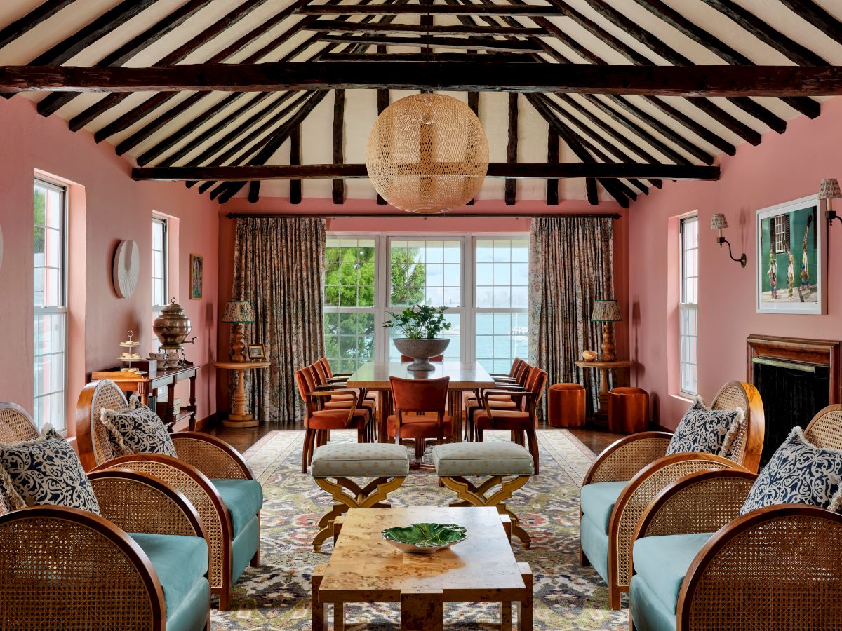 A cozy, pink-painted living room with wooden beams, light blue chairs, and a central coffee table, leading to a bright windowed area.