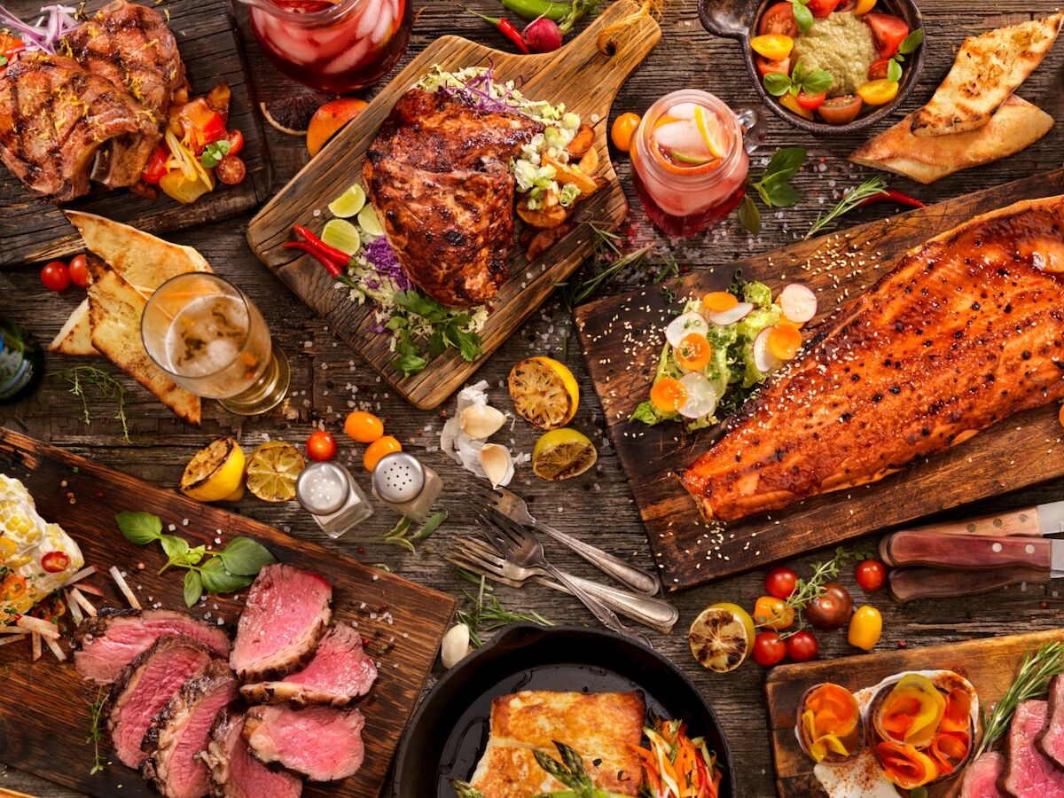 An assortment of grilled meats, bread, and fresh vegetables with herbs and sauces spread across a rustic wooden table.