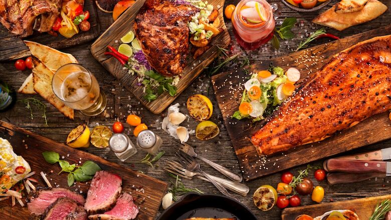 An assortment of grilled meats, bread, and fresh vegetables with herbs and sauces spread across a rustic wooden table.