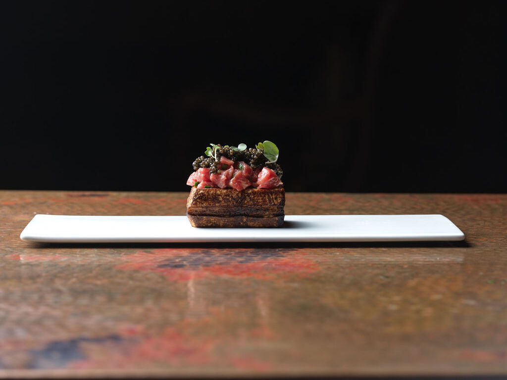 A mini tart on a white plate topped with diced meat, herbs, and seeds, served on a wooden table under a dark backdrop.