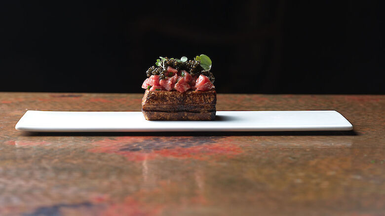 A mini tart on a white plate topped with diced meat, herbs, and seeds, served on a wooden table under a dark backdrop.