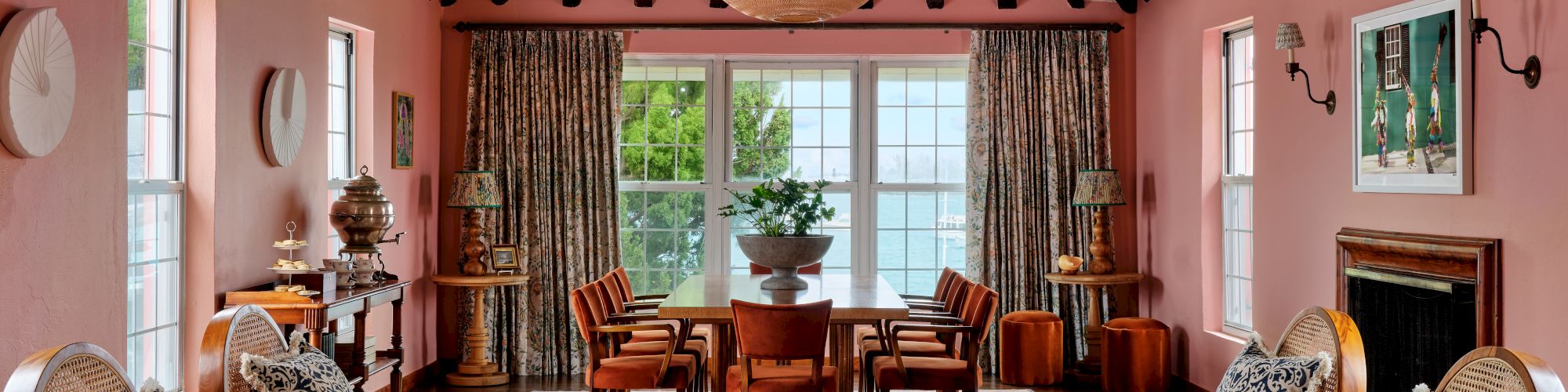A spacious living room with pink walls, exposed beams, wicker furniture, decorative items, large windows, and a central chandelier, ending the sentence.