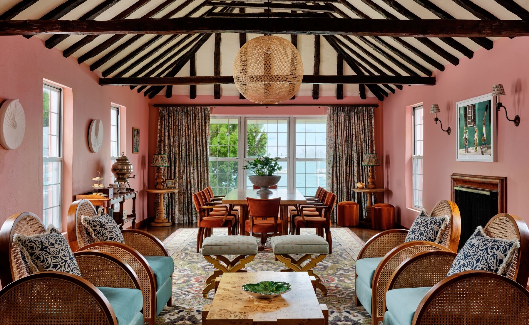A elegant living room with pink walls, wicker chairs, wooden beams, and a central coffee table beneath a round pendant light.