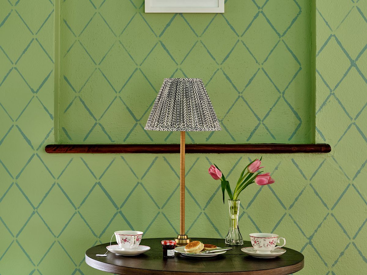 A small round table with a lamp, tea set, and vase of tulips, set against a green patterned wall with artwork.