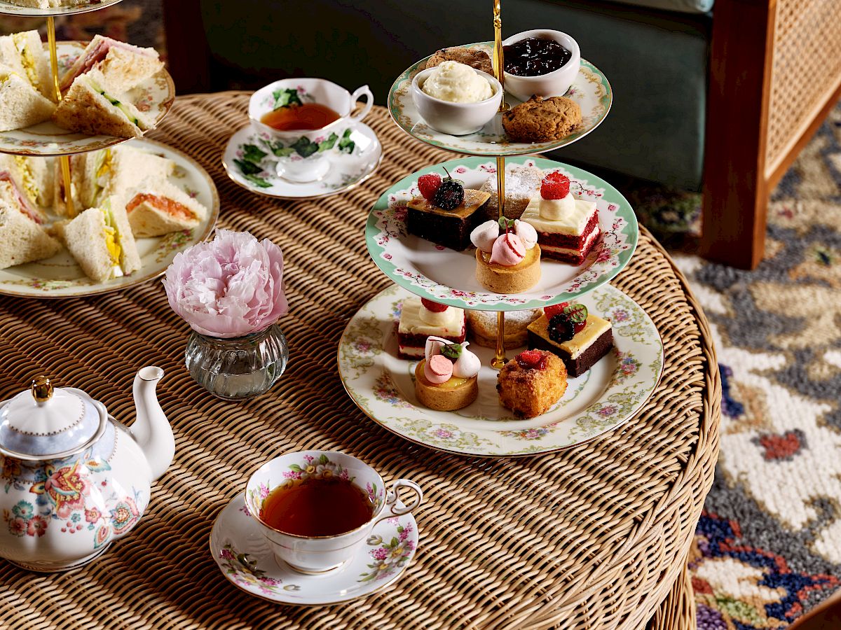 The image shows an elegant afternoon tea setup with a variety of pastries, sandwiches, a teapot, and teacups on a wicker table in a cozy seating area.