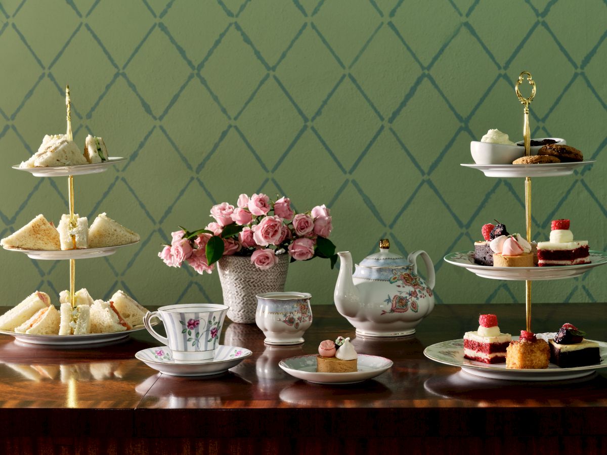 The image features a tea set arrangement with sandwiches, pastries on tiered trays, a teapot, cups, and a floral centerpiece on a wooden table.