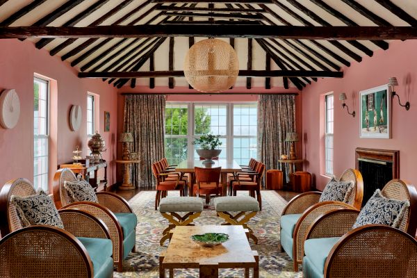 A cozy living room with a pink color scheme, featuring wooden beams, wicker furniture, a large central table, and windows with lush outside views.