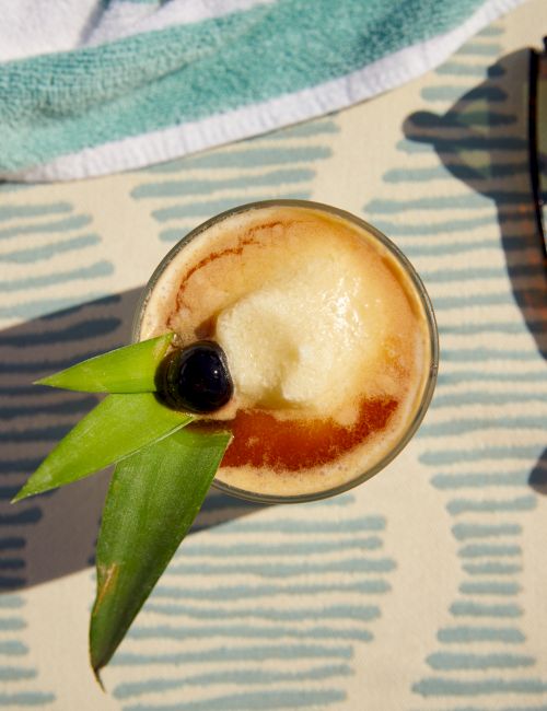 A tropical drink garnished with a cherry and pineapple leaves, sunglasses, and a towel are placed on a patterned surface, casting shadows.