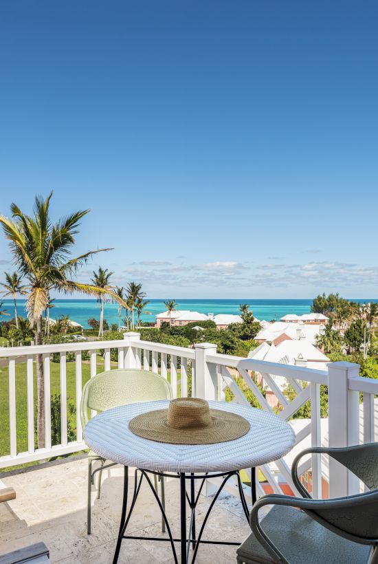 A scenic view from a balcony with a table and two chairs, overlooking a tropical landscape with palm trees and a clear blue ocean in the background.