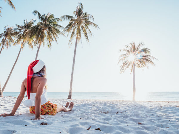 A woman in a red Santa hat relaxes on a sandy beach beneath palm trees by the ocean, enjoying sunny holiday vibes.