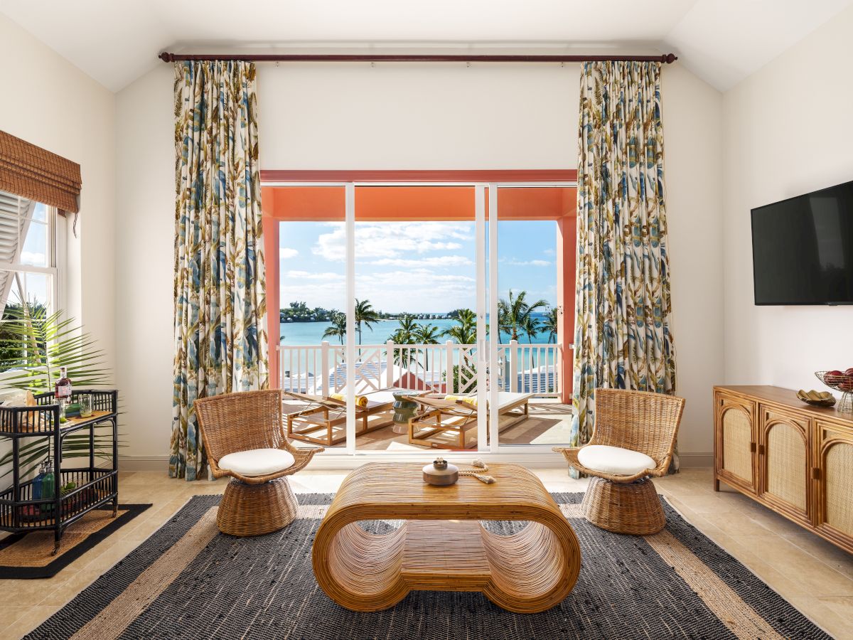 A living room with wicker furniture, patterned curtains, a woven rug, and a large window leading to a balcony with a view of the ocean and palm trees.
