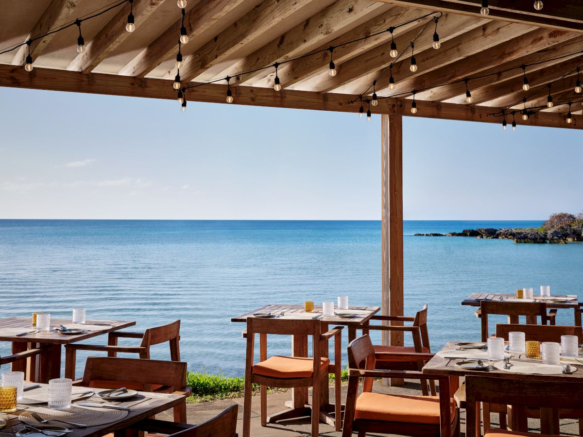 An outdoor restaurant with wooden tables and chairs under a pergola, overlooking a calm sea with a clear blue sky.